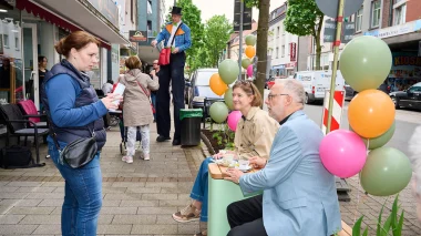 Hertens Bürgermeister Matthias Müller sitzt zwischen bunten Ballons auf dem Parklet auf der Bahnhofstraße vor der Eisdiele, Stelzenläufer "Rainer Blödsinn" ist im Hintergrund zu sehen